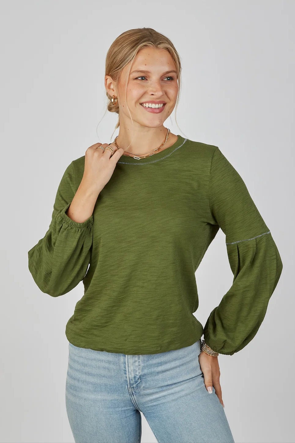 Woman wearing a green long-sleeve top and blue jeans on a plain background