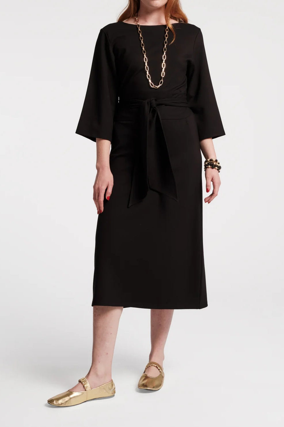 A woman modeling a black midi dress with three-quarter sleeves, a boat neckline, and a matching fabric tie belt at the waist. She is styled with a chunky gold chain necklace, bracelets, and gold flat shoes against a plain light gray studio background.