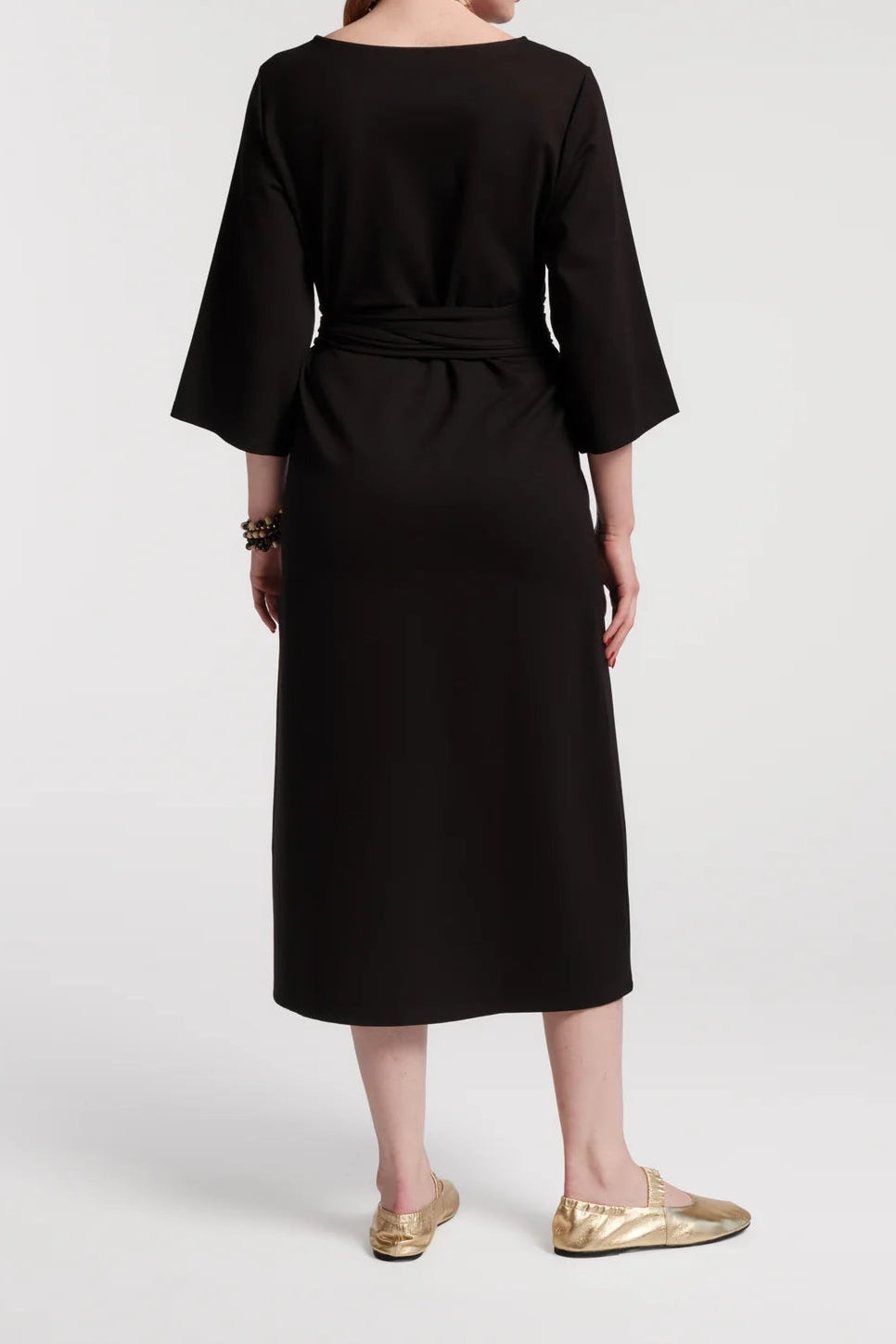 A woman with red hair is shown from the back wearing a black midi dress with three-quarter sleeves and a matching fabric tie belt at the waist. She is wearing gold metallic flats against a light gray studio background.