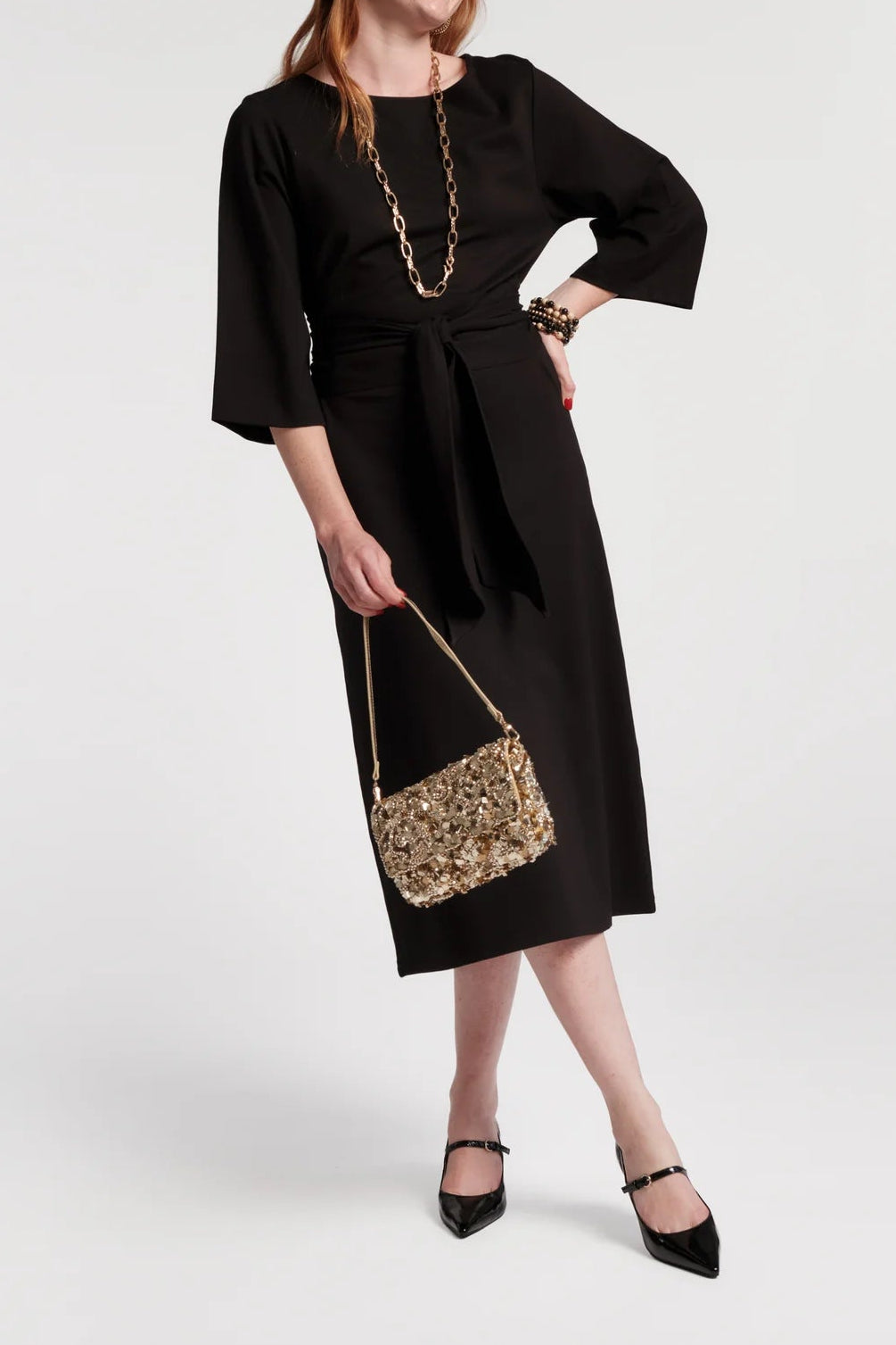 A woman modeling a black midi dress with three-quarter sleeves and a self-tie waist, styled with a chunky gold chain necklace, a gold-embellished shoulder bag, and black pointed flats against a light gray studio background.