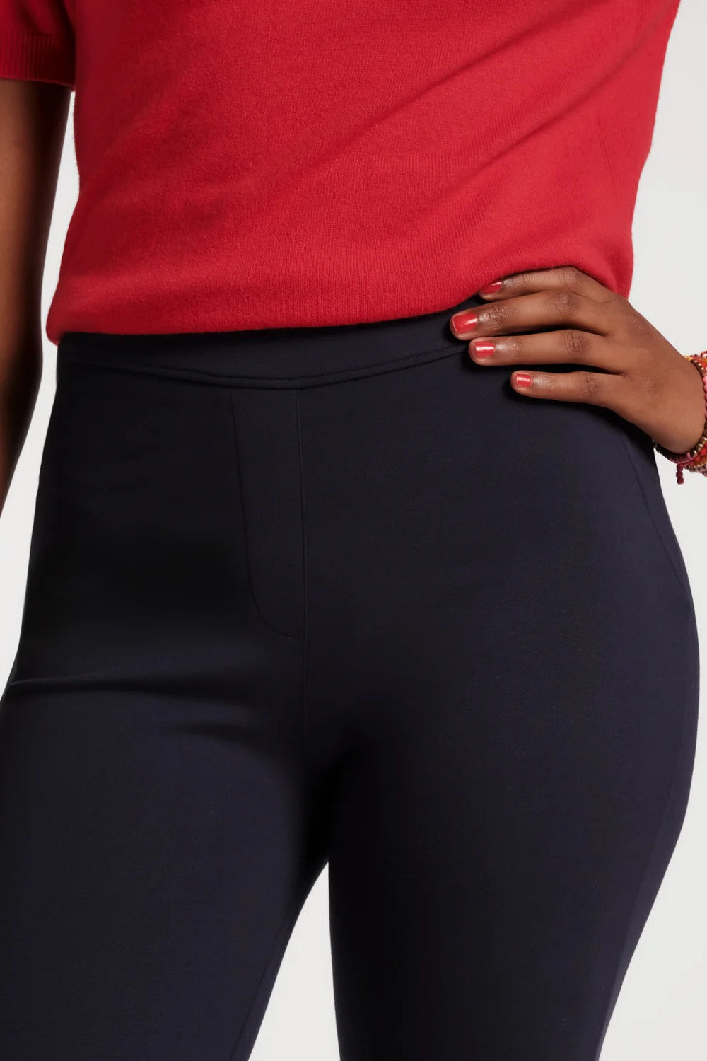 Close-up of a person wearing dark navy high-waisted leggings and a red short-sleeve top, with one hand resting on the hip against a light gray studio background.