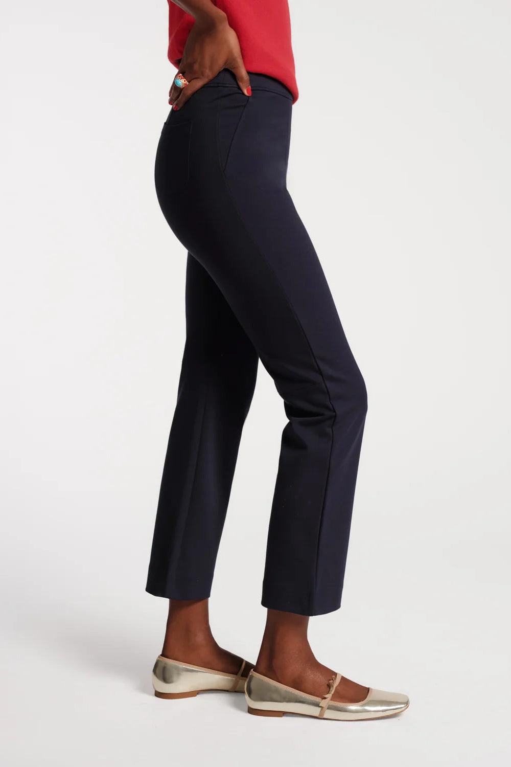 A side-profile view of a model wearing dark navy blue cropped trousers, paired with a red top and metallic gold flats, against a clean white studio background.