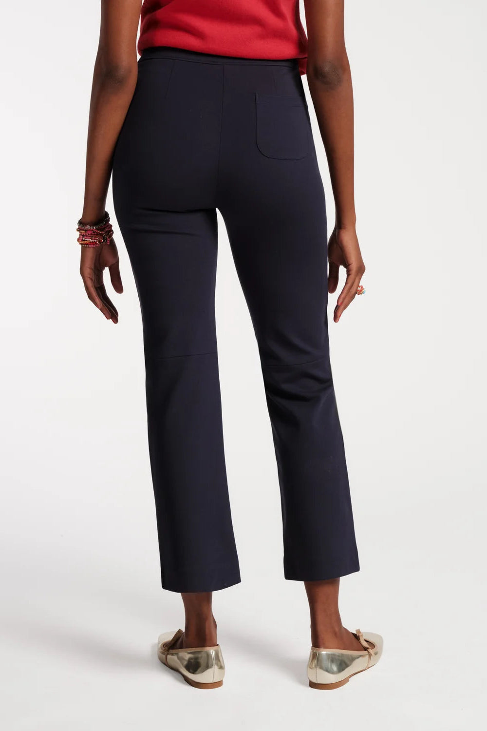 A model shown from the back wearing dark navy cropped trousers, a red top, and metallic flat shoes against a plain white studio background.