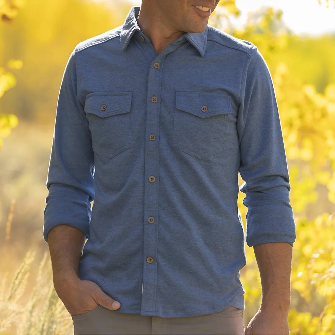 A man wearing a blue long-sleeve button-up shirt with two chest pockets and a brown cowboy hat, standing outdoors in a field of bright yellow autumn leaves.