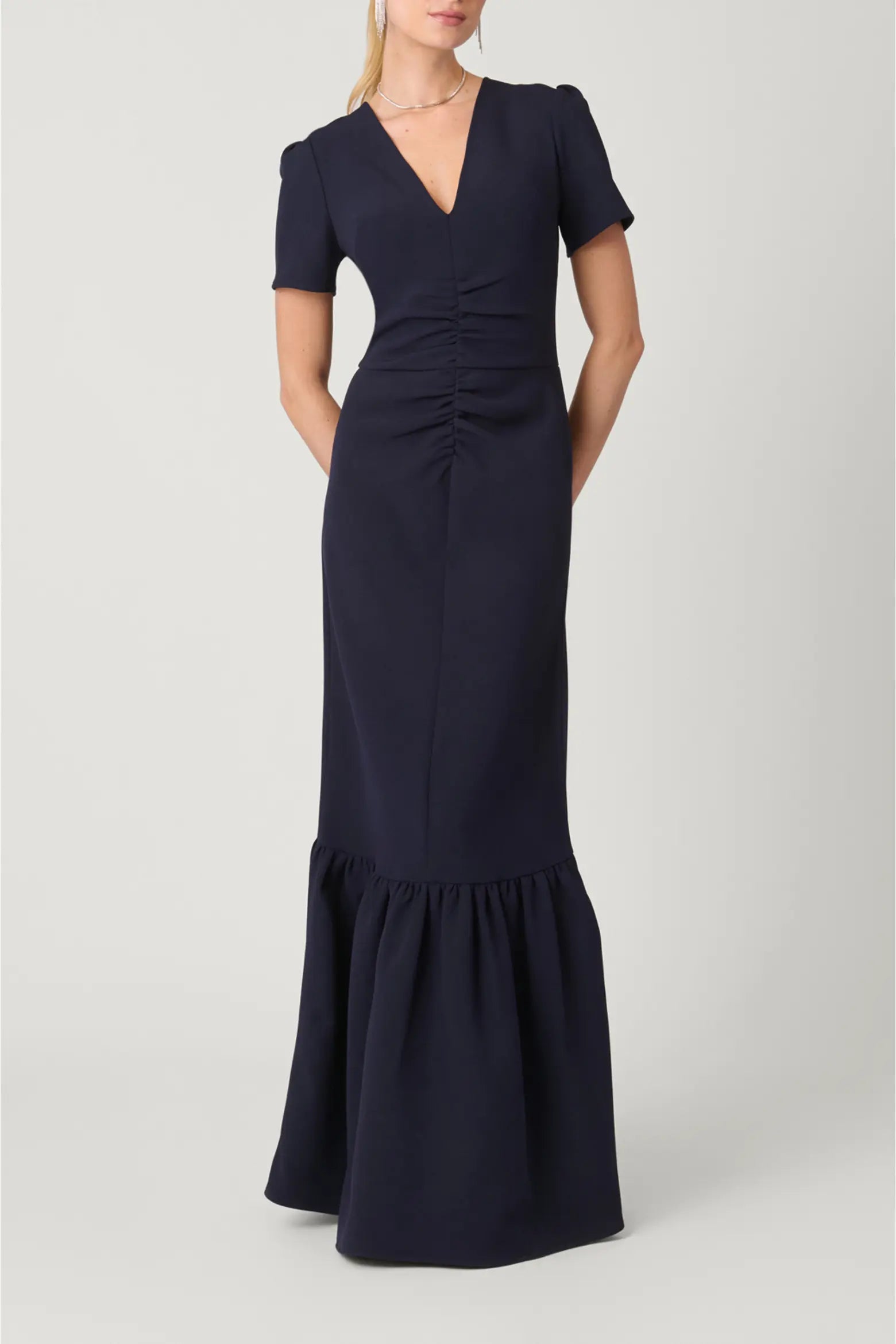 A woman modeling a navy blue floor-length gown with short sleeves, a deep V-neckline, gathered ruching at the waist, and a flared ruffled hem, photographed against a light gray studio background.