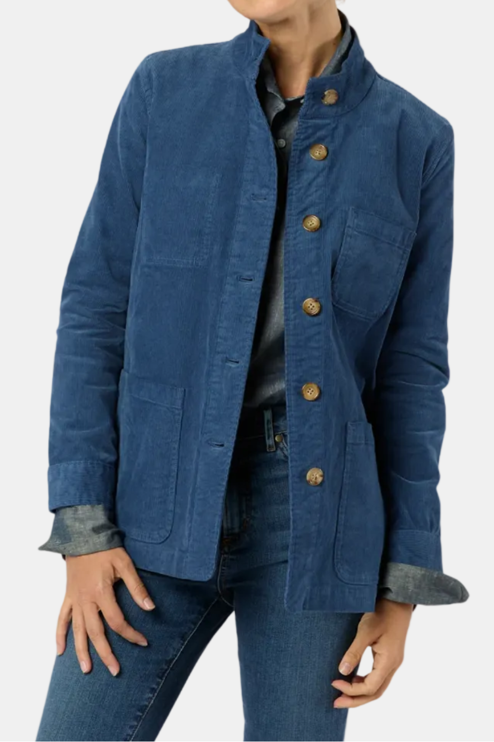A woman wearing a blue corduroy jacket with brown buttons, layered over a dark shirt and paired with blue jeans, photographed against a light gray studio background.