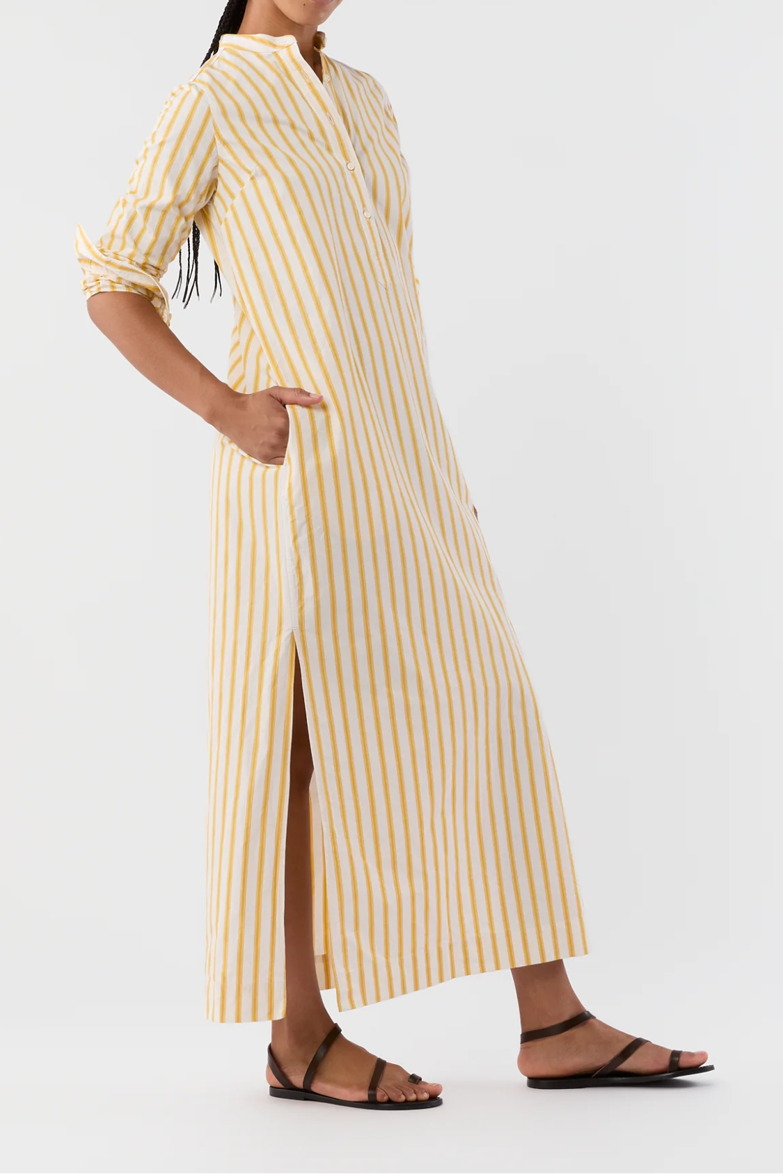 A woman modeling a long yellow and white vertically striped midi dress with a stand collar, three-quarter sleeves, and a front slit, paired with black strappy flat sandals against a plain white studio background.