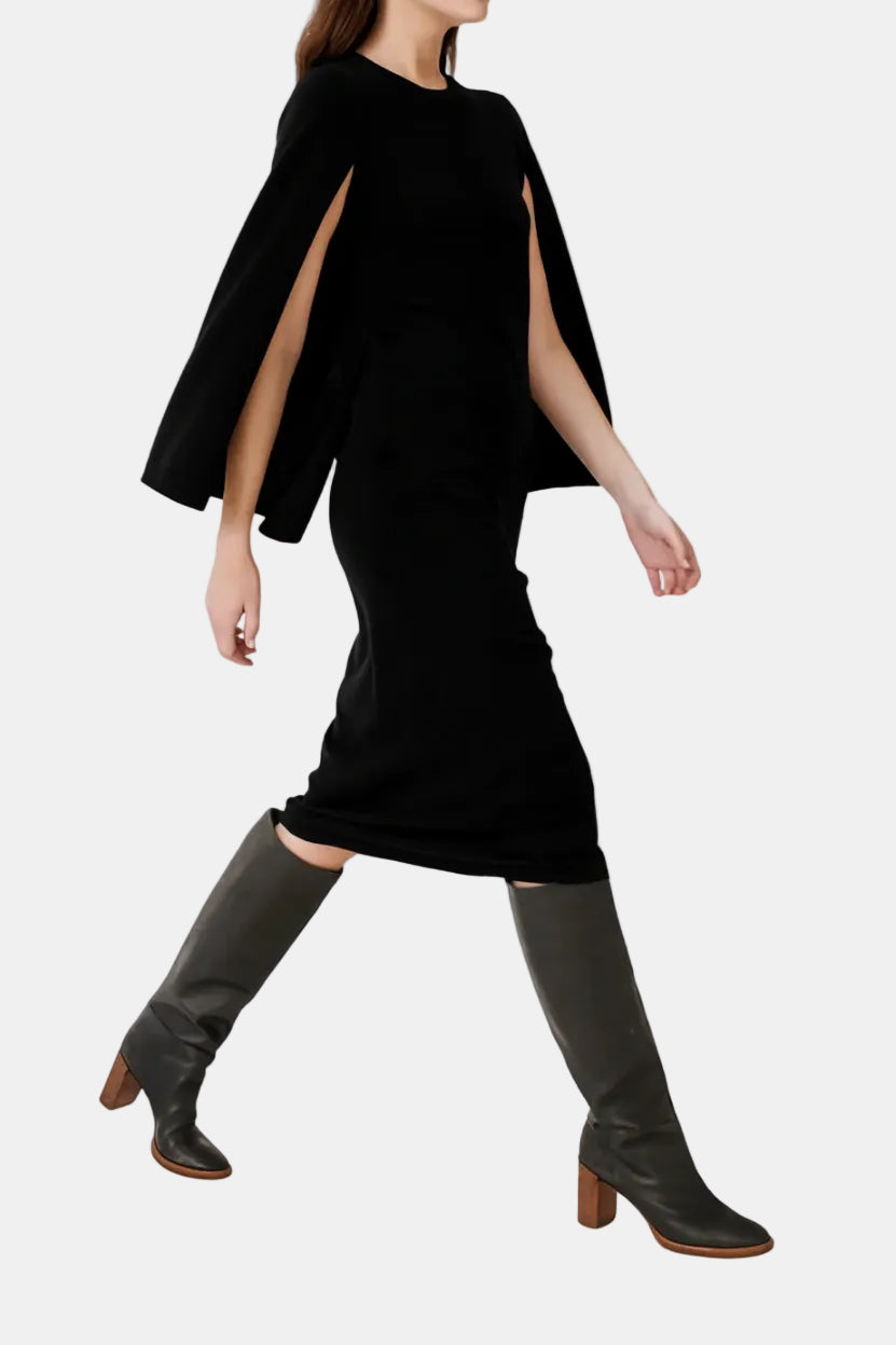A woman modeling a black long-sleeve dress with dramatic cape-like sleeves and side slits, paired with dark gray knee-high boots, against a plain white background.