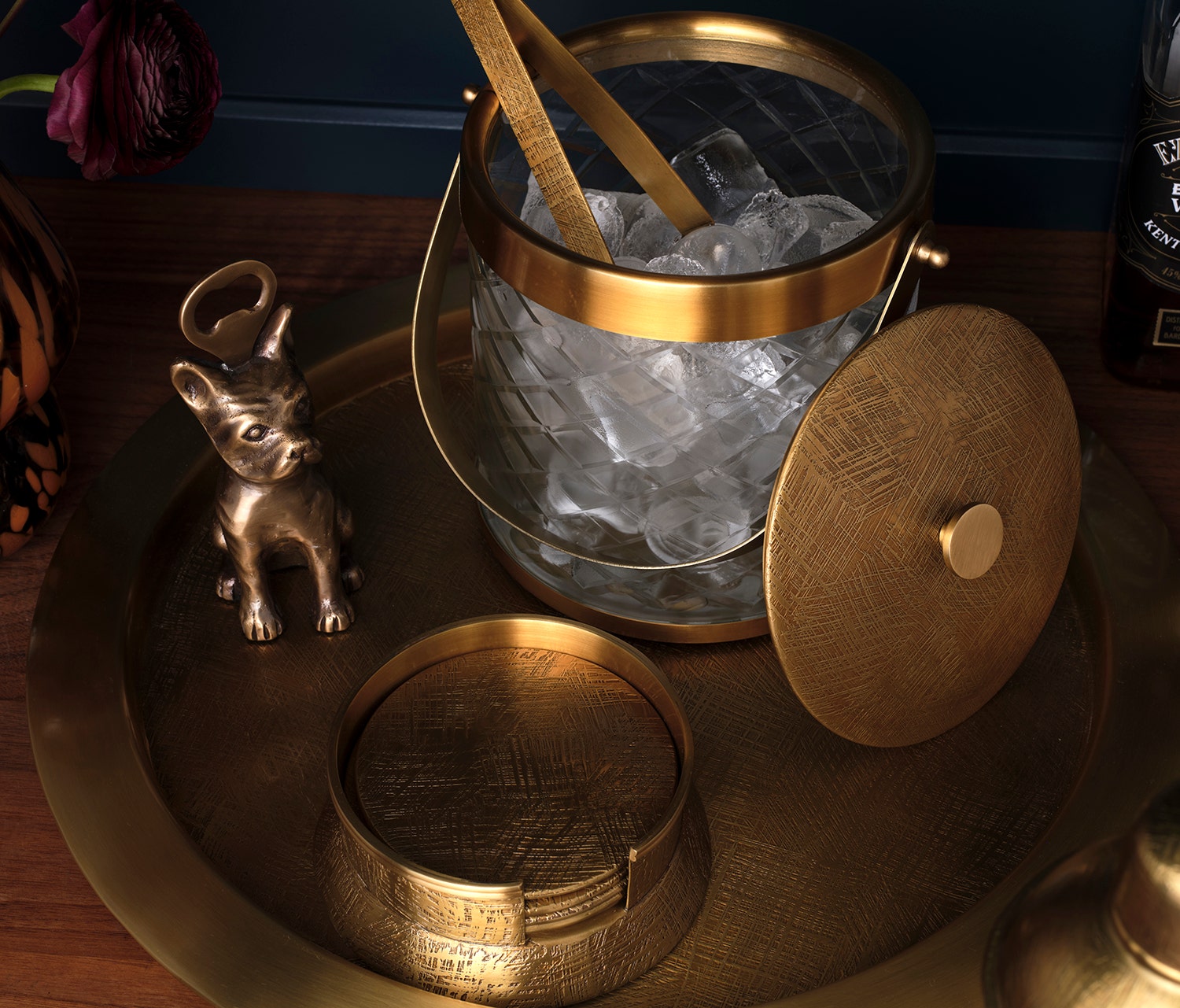 Gold ice bucket with ice and a wooden spoon on a wooden tray, with a small decorative dog figurine nearby.