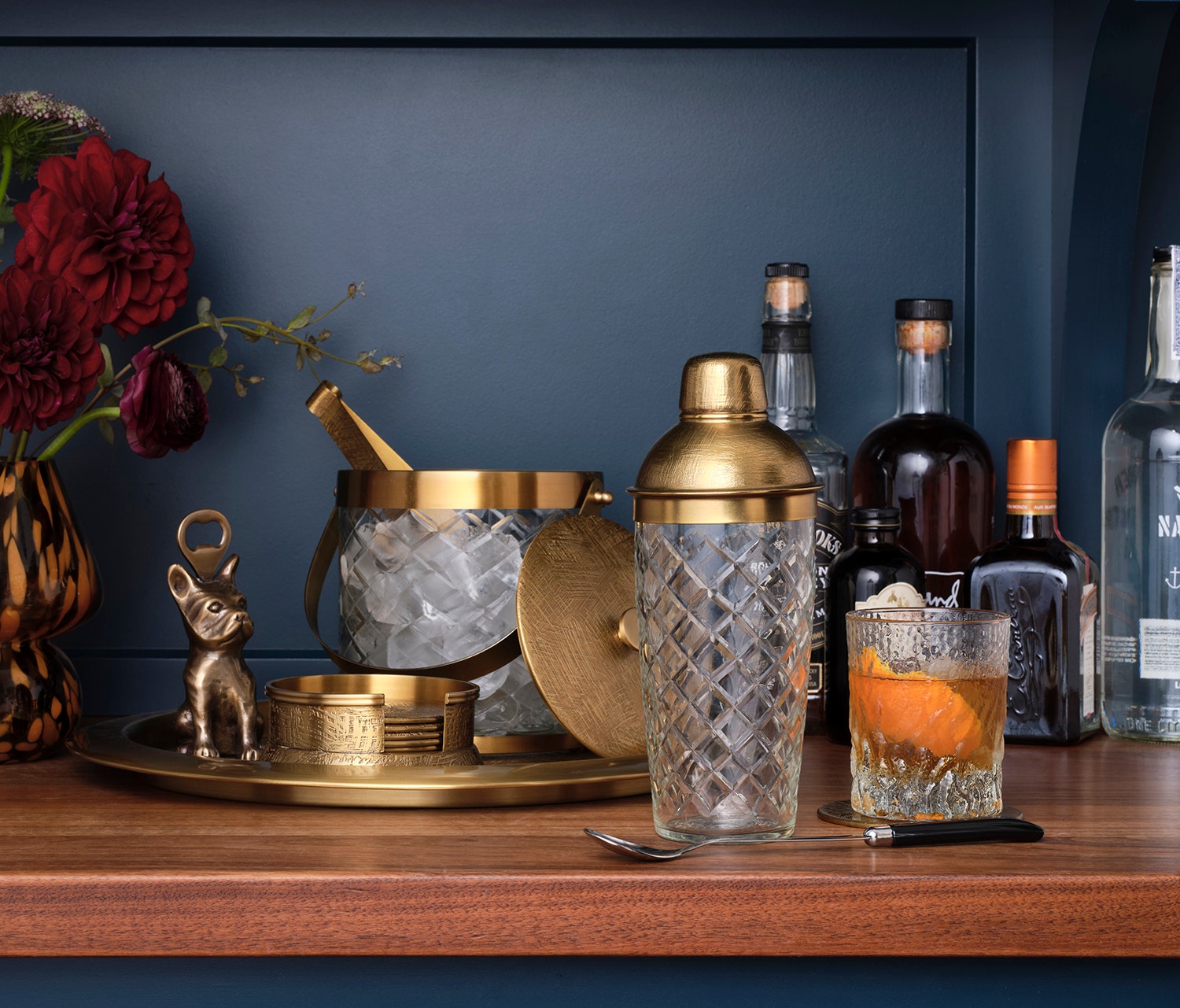 Bartending setup with cocktail shaker, bottles, and glass on a wooden surface against a dark blue wall.