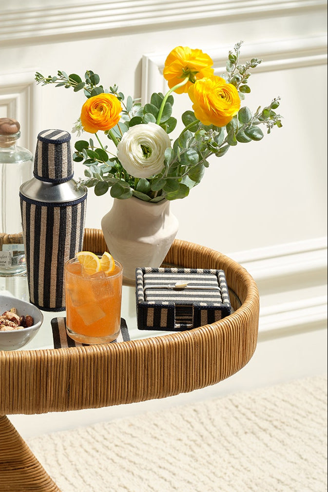 A round woven rattan bar cart styled with cocktail accessories, including striped tumblers, a shaker, a bottle, a small bowl of olives, a drink with a lemon slice, and a vase of yellow and white flowers, set against a white paneled wall.