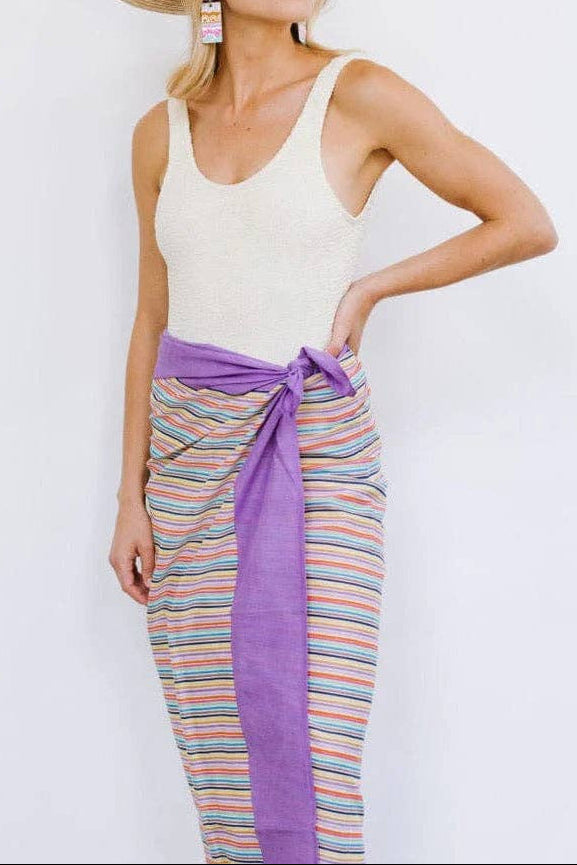 Woman wearing a white tank top and colorful striped sarong with a straw hat on a light background