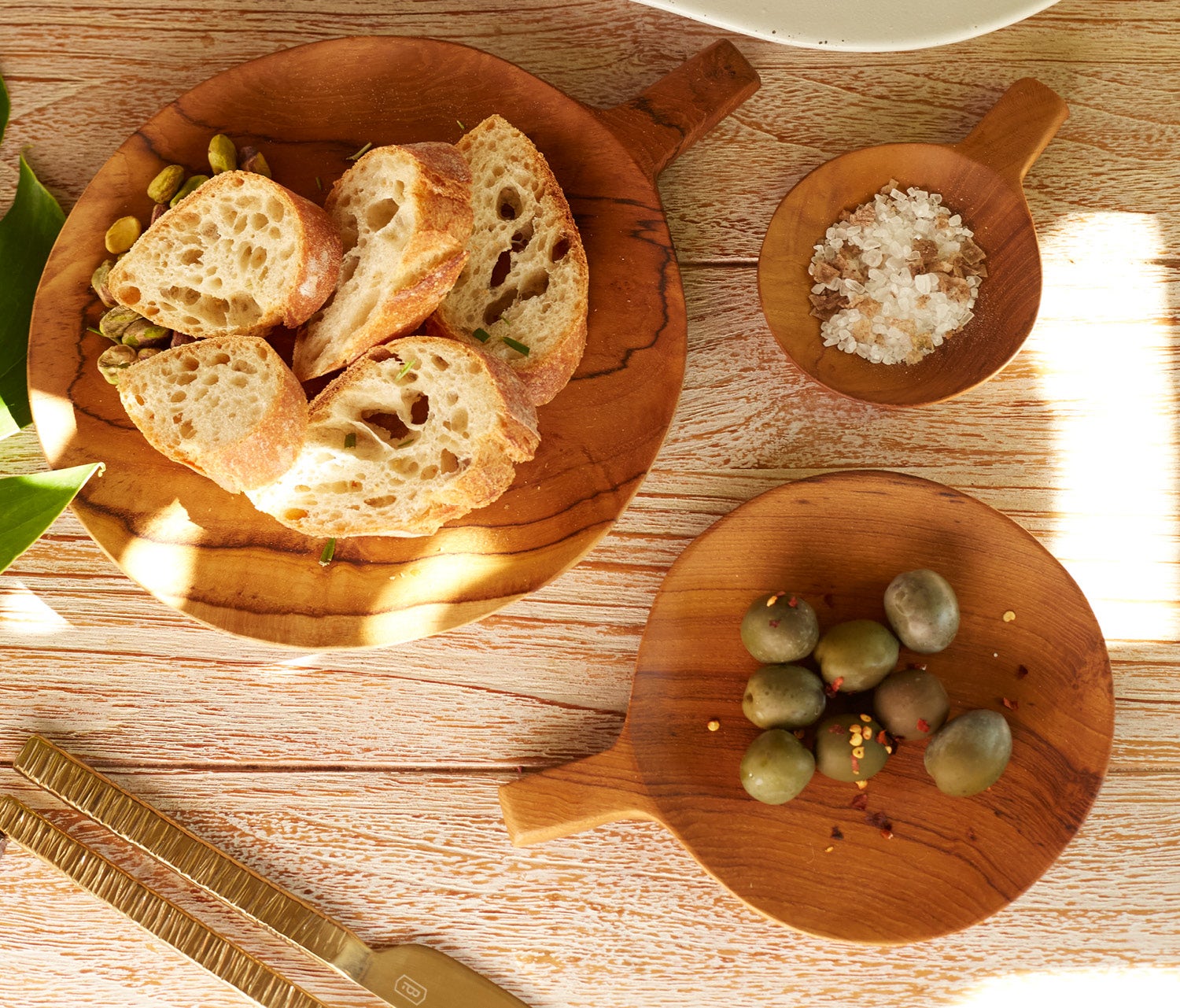 Three round wooden serving boards with handles are arranged on a light wood tabletop. One board holds sliced artisan bread with green beans, one holds coarse salt, and one holds green olives.