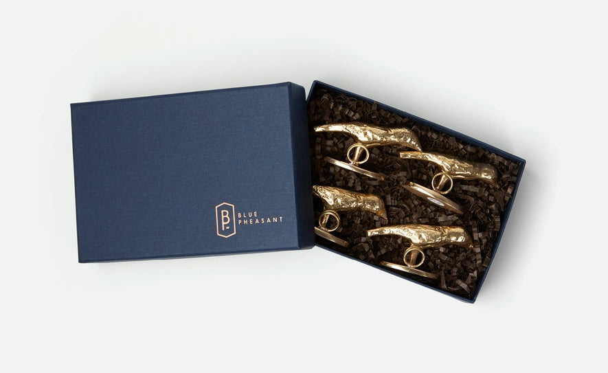 A navy blue jewelry gift box, partially open to reveal five gold-tone ring-shaped accessories nestled in dark crinkle paper. The closed lid on the left features a gold logo that reads 'BLUE PLEASANT'.