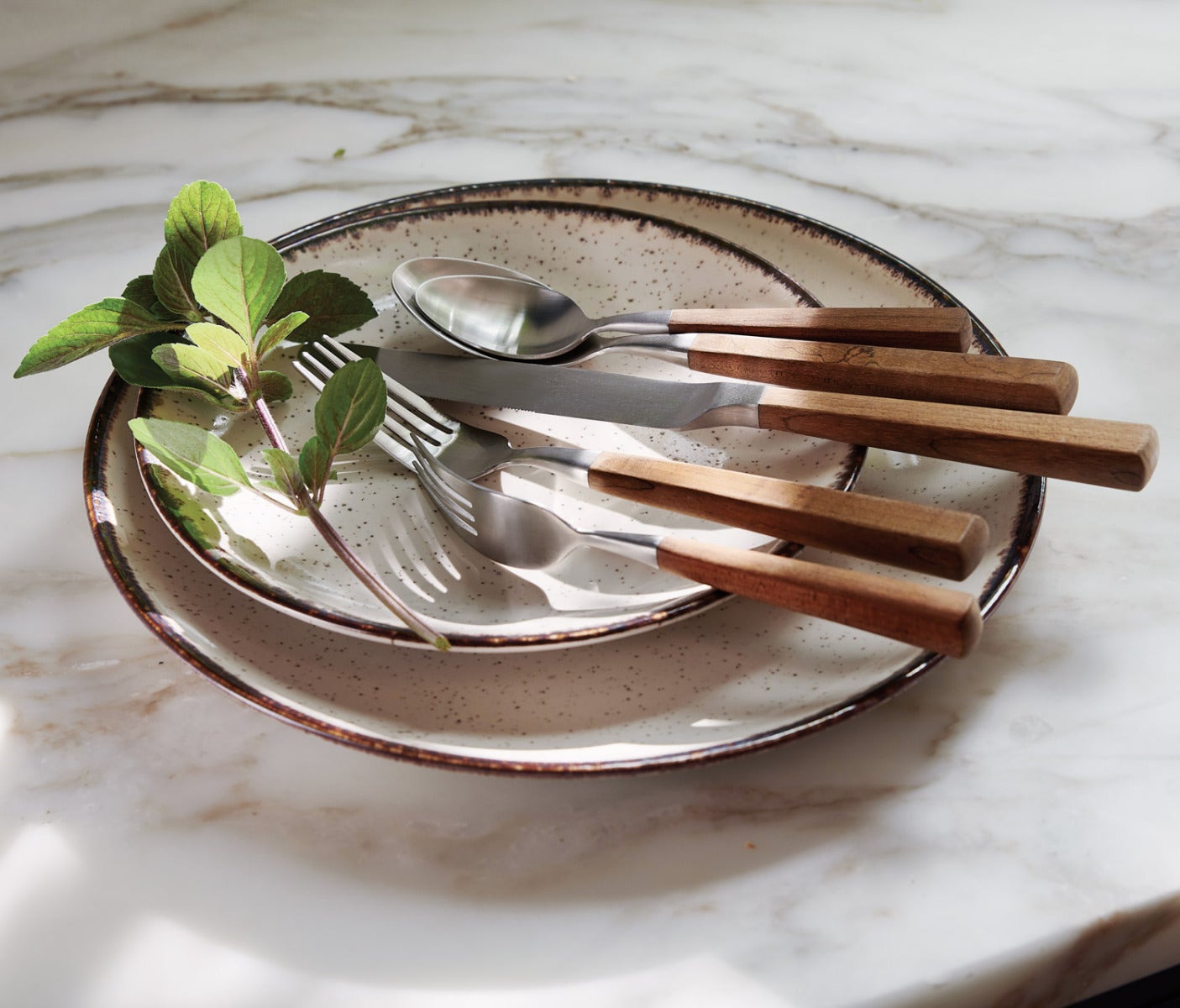 A stack of speckled beige ceramic plates with dark brown rims, styled with silver metal cutlery featuring natural wood handles, alongside green herb leaves on a white marble surface.