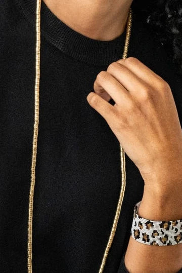 A close-up of a person wearing a wide cuff bracelet with a black, white, and brown geometric pattern, paired with a black sweater and gold chain.