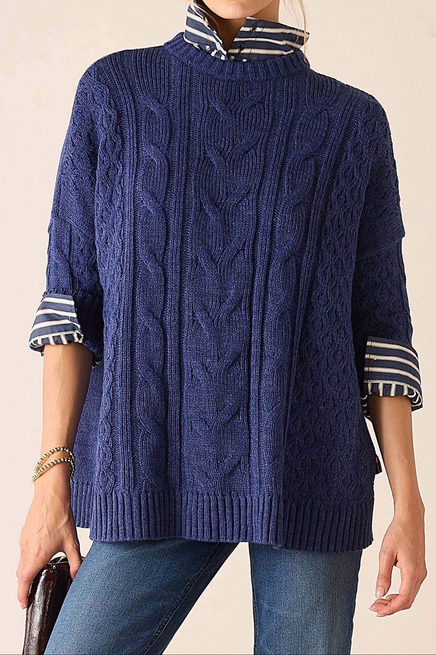 A woman wearing a navy blue cable-knit mock-neck sweater with striped knit cuffs, layered over a matching striped mock-neck shirt, paired with blue jeans against a plain beige background.
