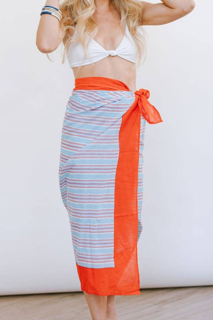 Woman wearing a white bikini top and a striped sarong with orange sides on a plain background
