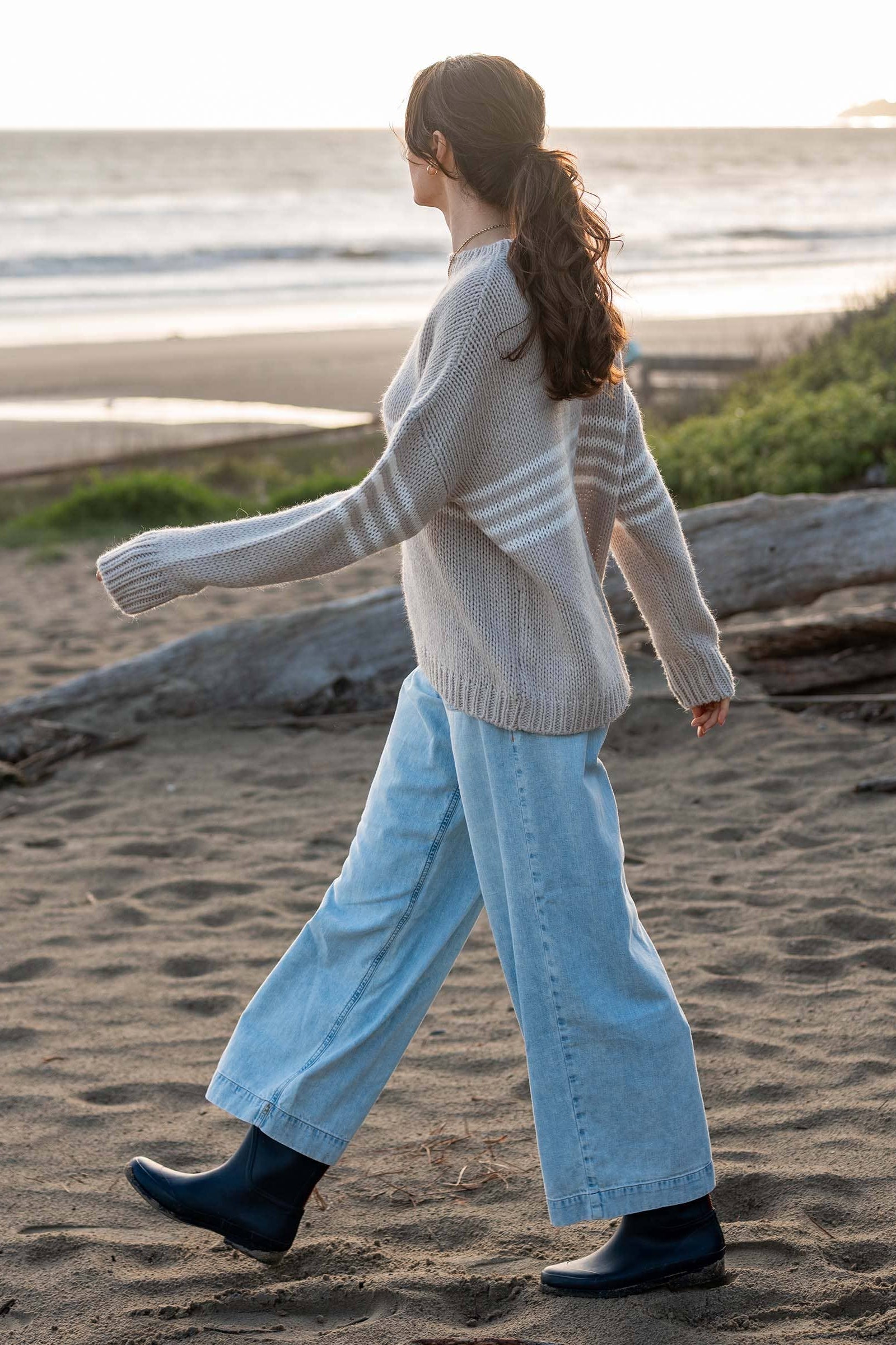 Woman walking on a beach wearing a light sweater and blue jeans.