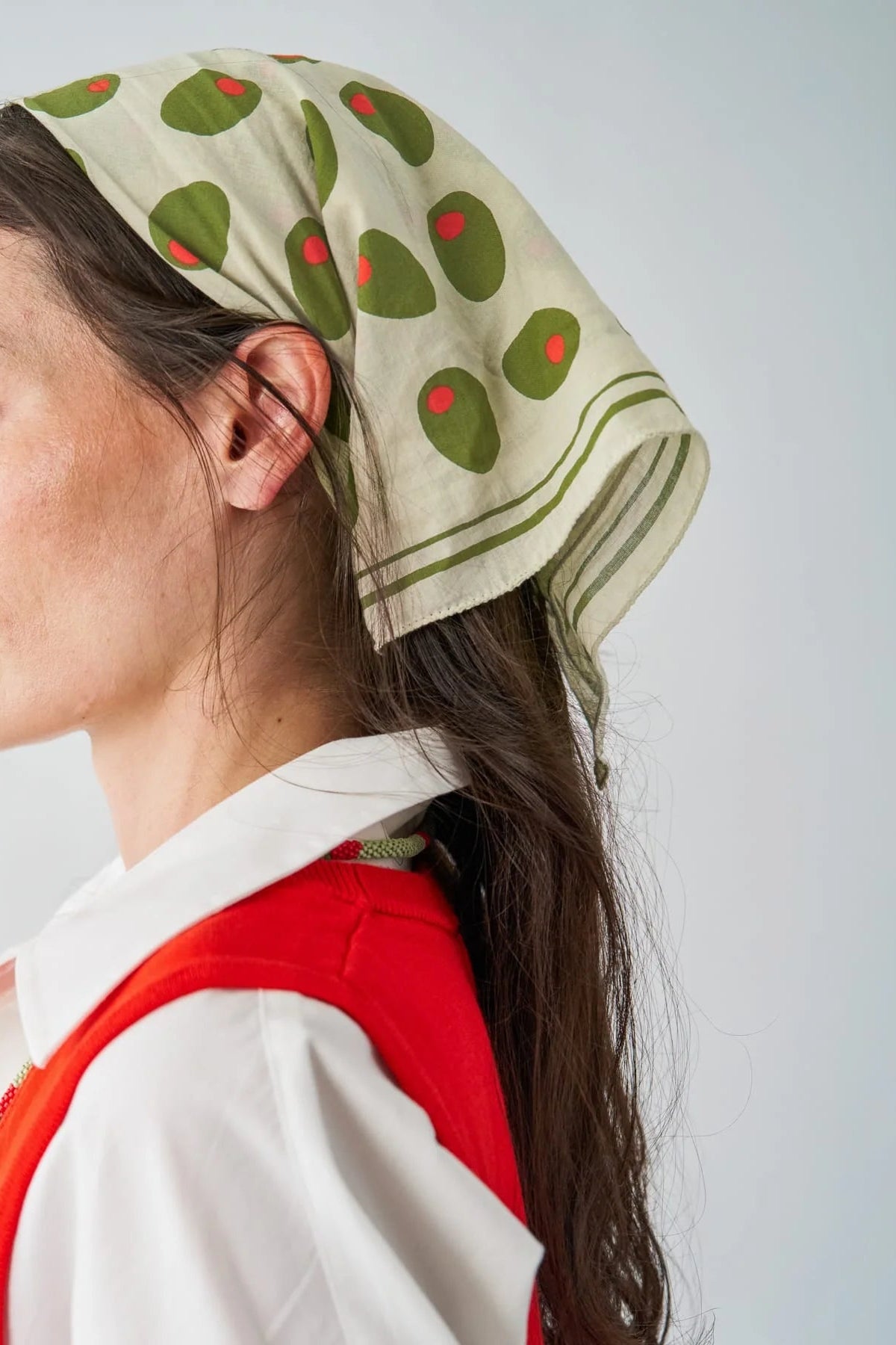 A woman in profile wearing a cream-colored headscarf with green and red patterned dots, paired with a red vest and white shirt, against a light gray background.