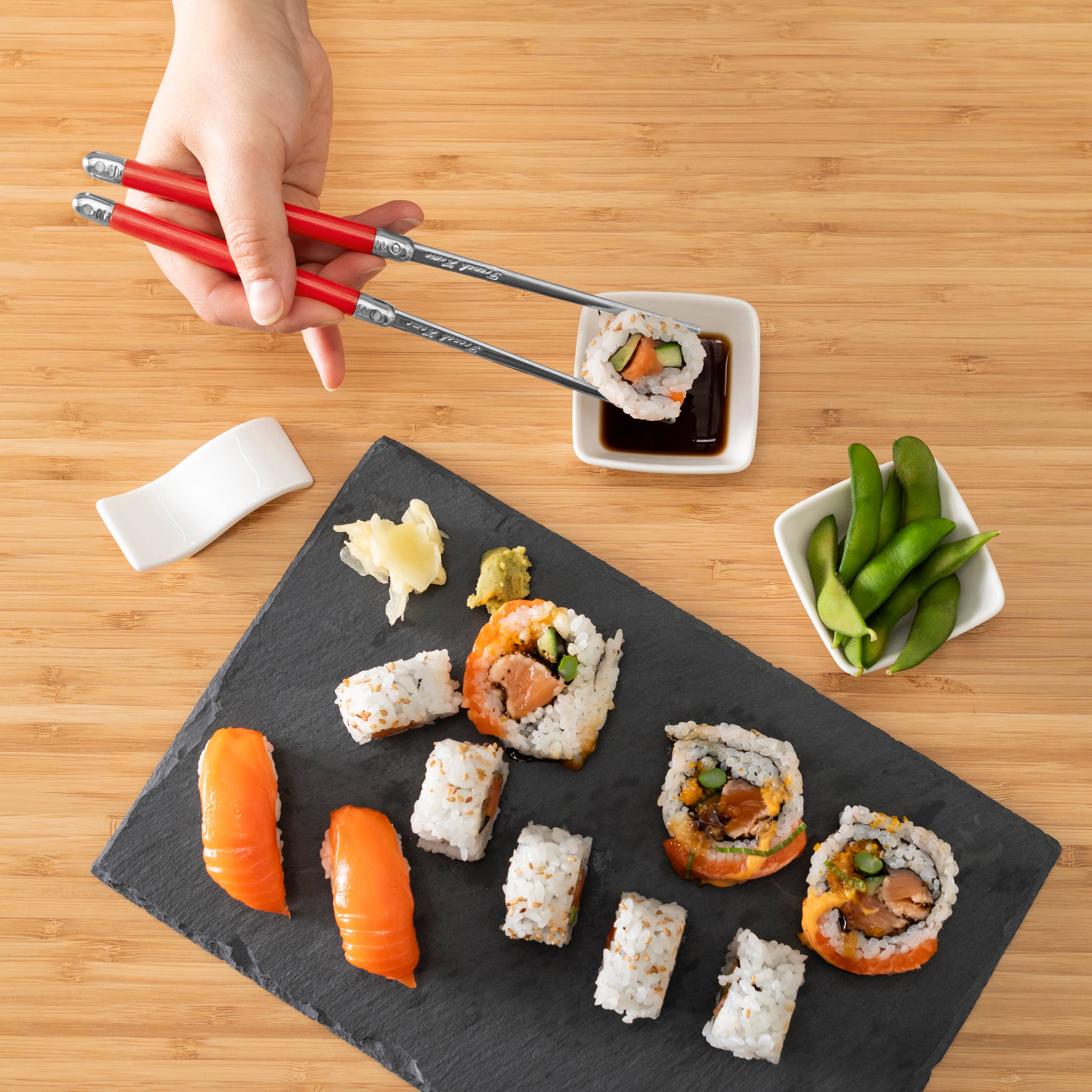 Sushi platter with assorted sushi rolls on a black slate, a hand using chopsticks, and small bowls of wasabi and green onions on a wooden table.