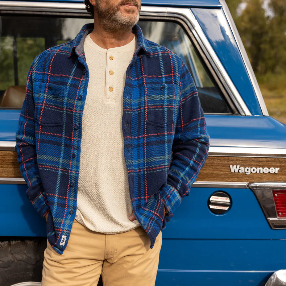 Man in a blue plaid shirt standing next to a blue Jeep Wagoneer in a natural setting.