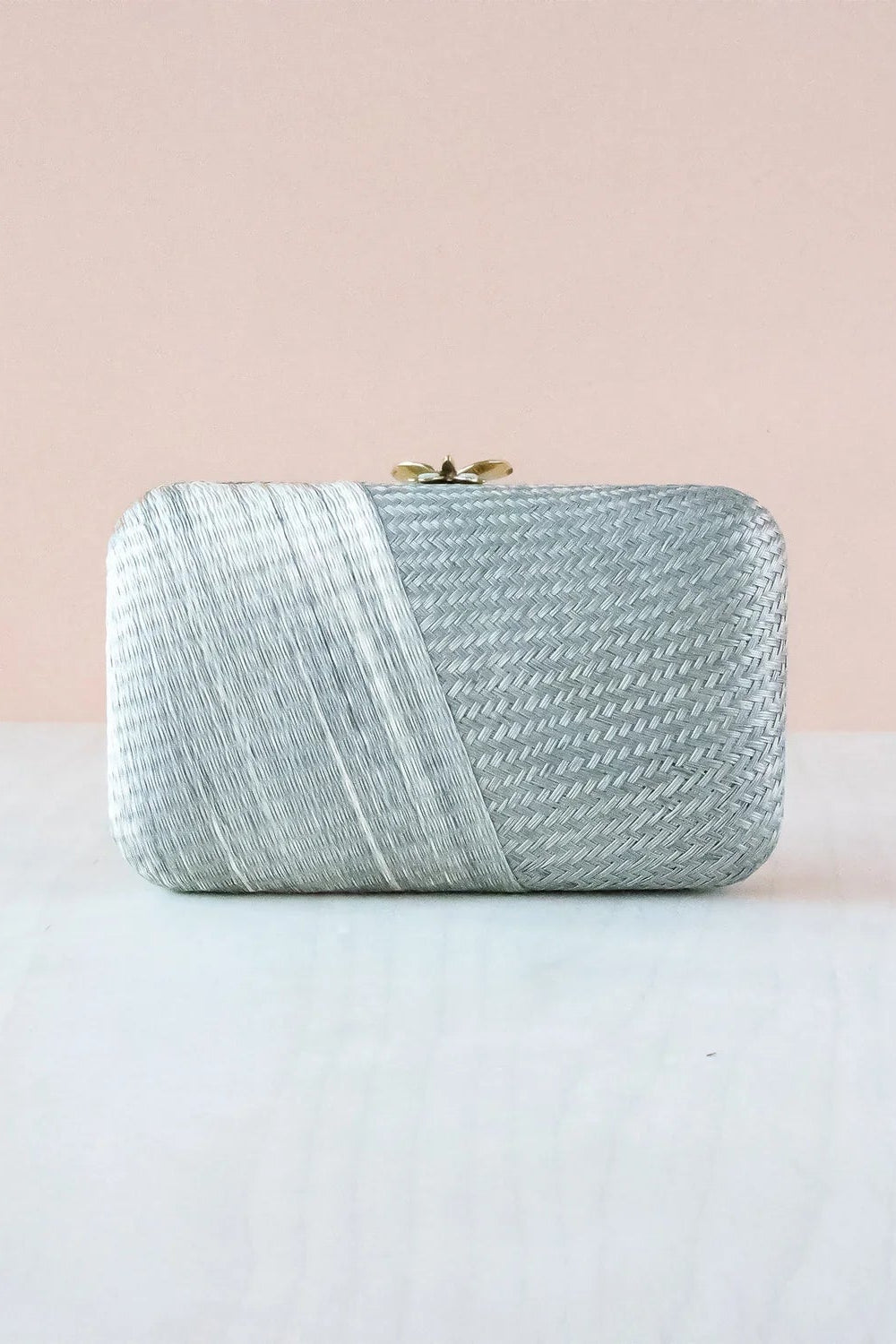 A silver textured clutch purse with a diagonal wrap design and a small gold clasp, resting on a white surface against a soft pink background.