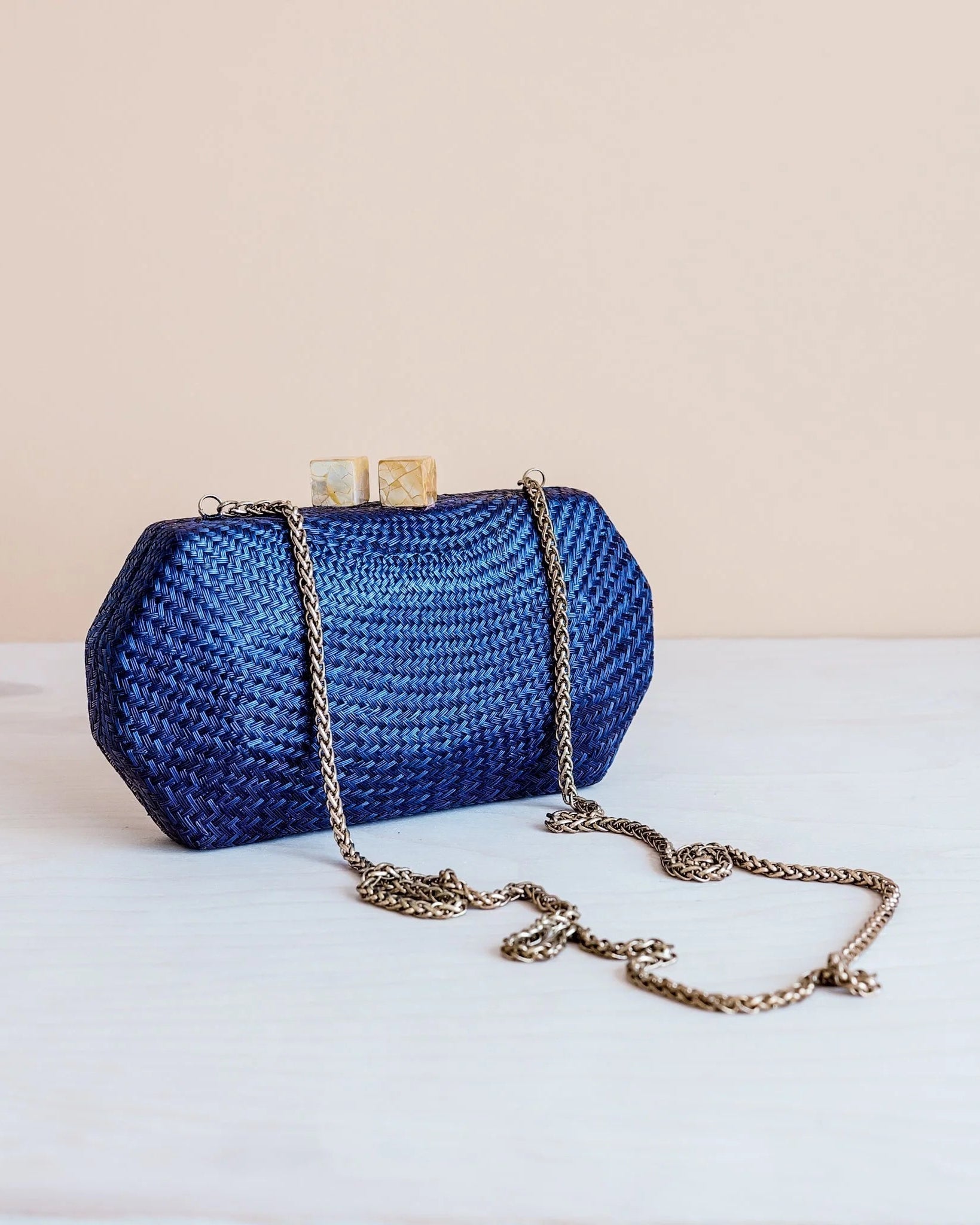 A blue woven clutch purse with a gold-tone chain strap, resting on a white surface against a light beige background.