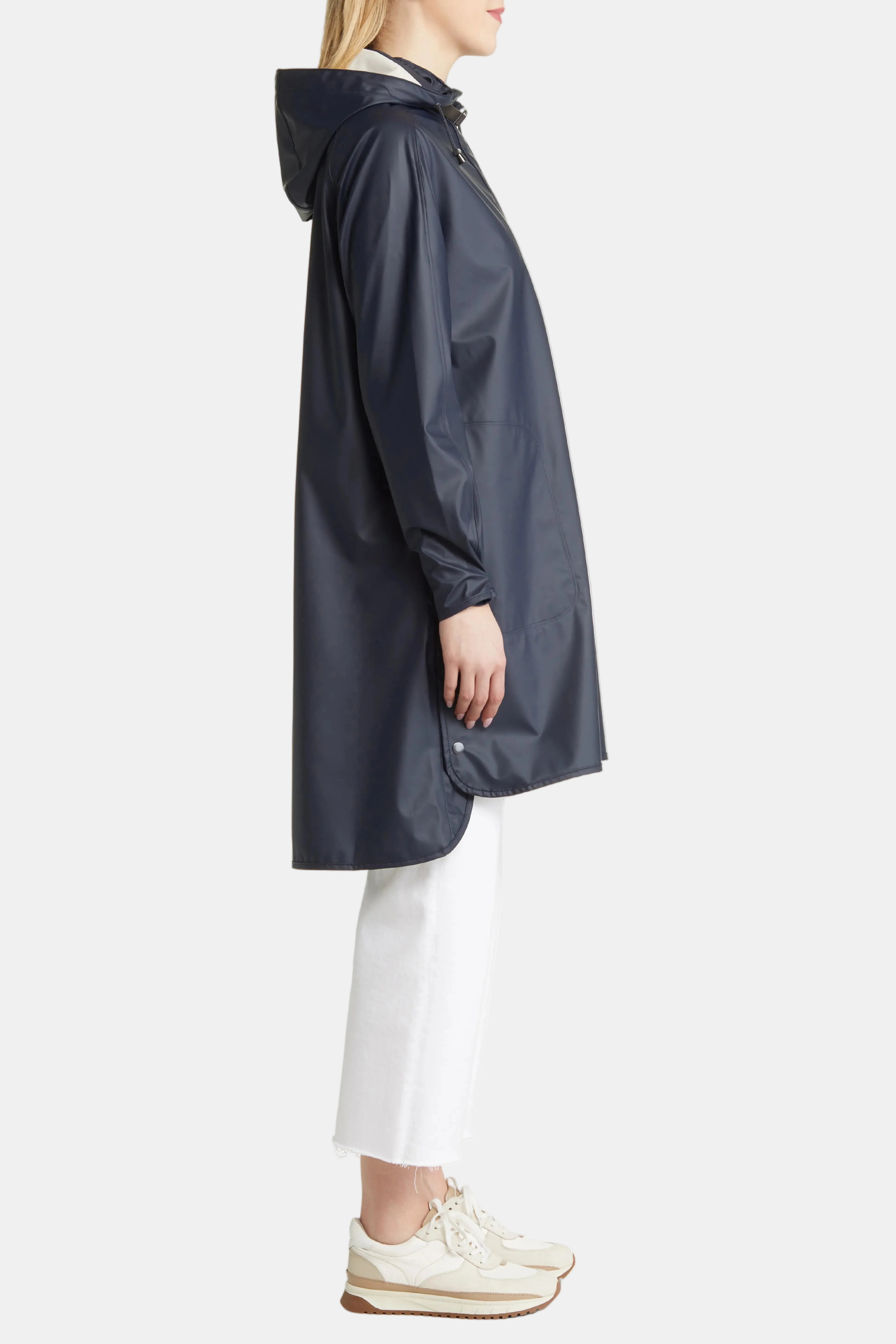A model shown from the side wearing a dark navy hooded raincoat, white cropped pants, and beige sneakers against a plain white background.