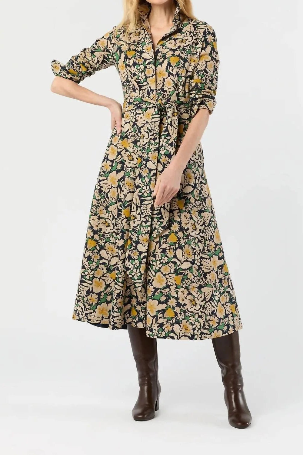 A woman models a mid-length button-front dress with three-quarter sleeves and a self-tie waist, featuring a dense floral and botanical print in green, yellow, and beige. She is wearing dark brown knee-high boots against a plain white studio background.