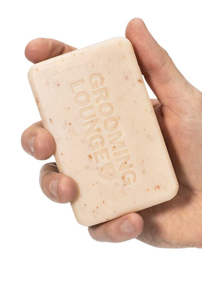 Hand holding a bar of soap with 'Grooming Lounge' branding on a white background