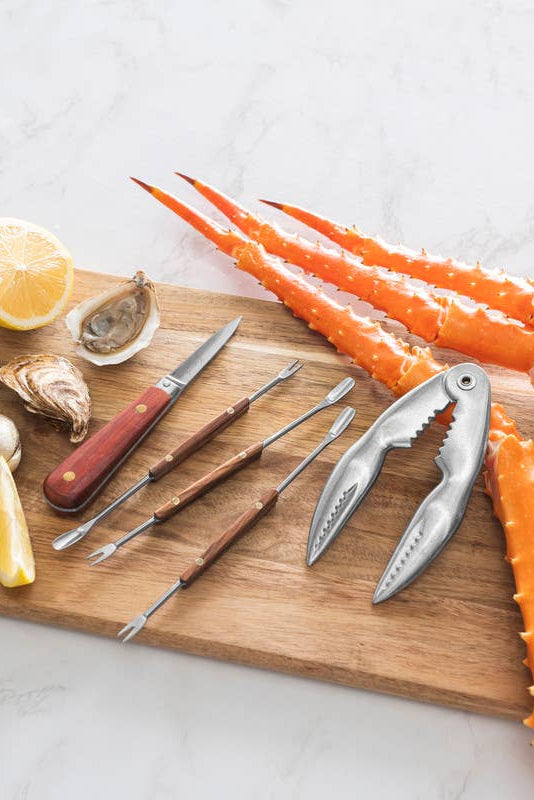 Wooden cutting board with seafood tools, clams, lemon, and crab legs on a white surface.