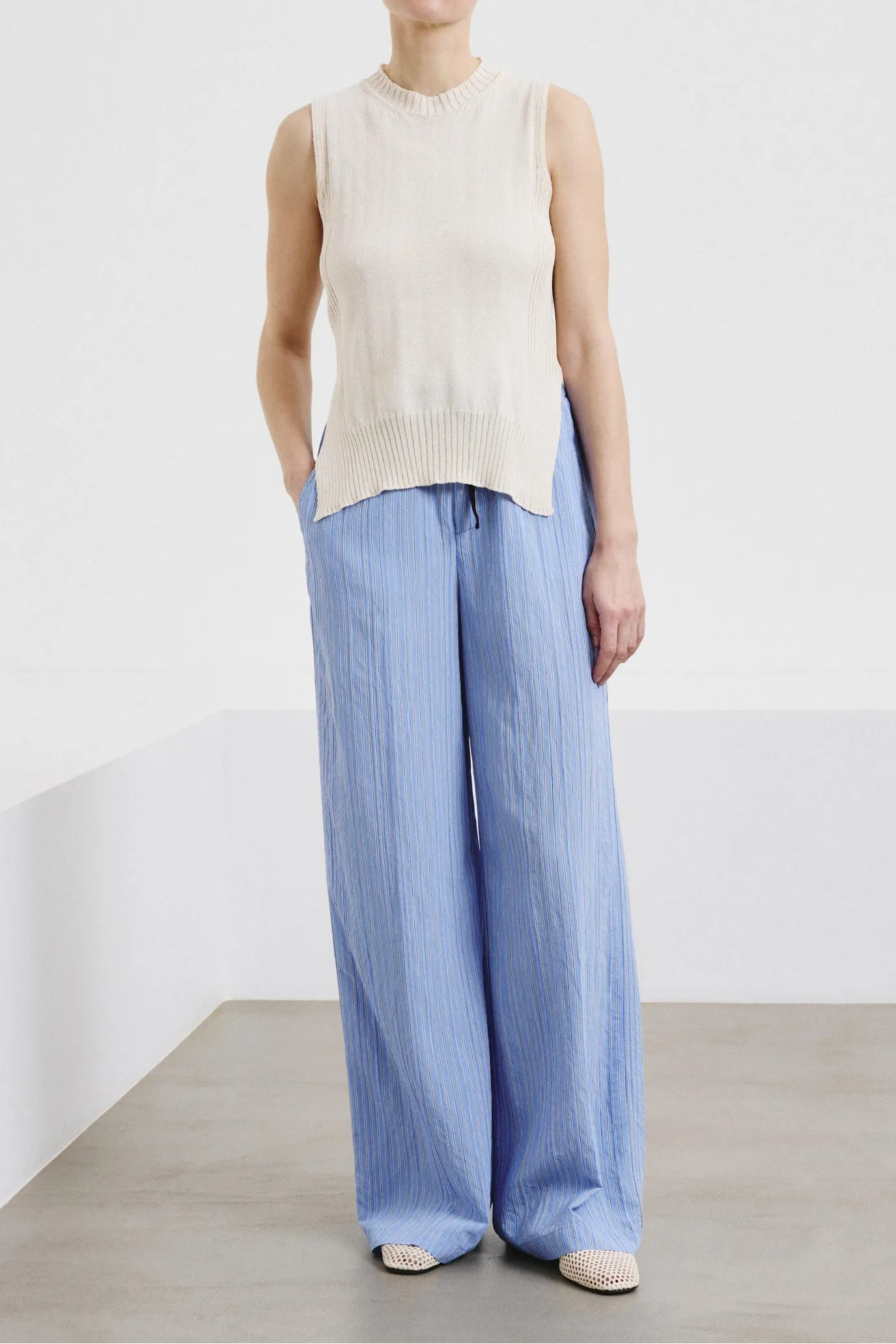 Woman wearing a sleeveless beige top and light blue pants against a white background