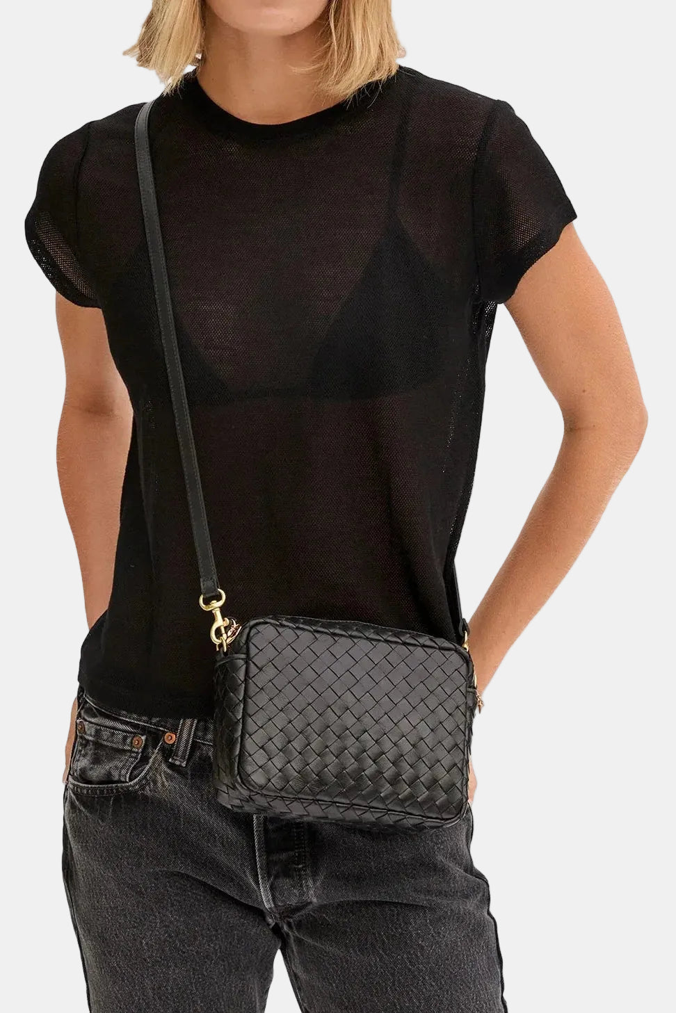 A woman wearing a black sheer short-sleeve top and dark gray jeans, carrying a black woven crossbody bag with a gold-tone logo clasp. The background is solid white.