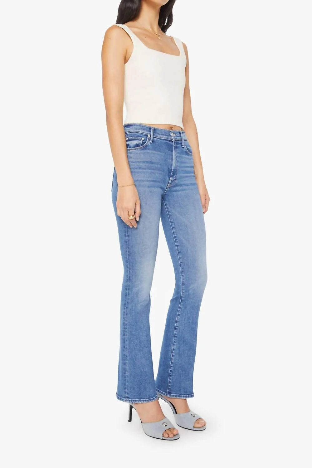 A model wearing light-wash blue flare jeans, a white ribbed tank top, and light blue heeled sandals, shown against a plain light gray studio background.