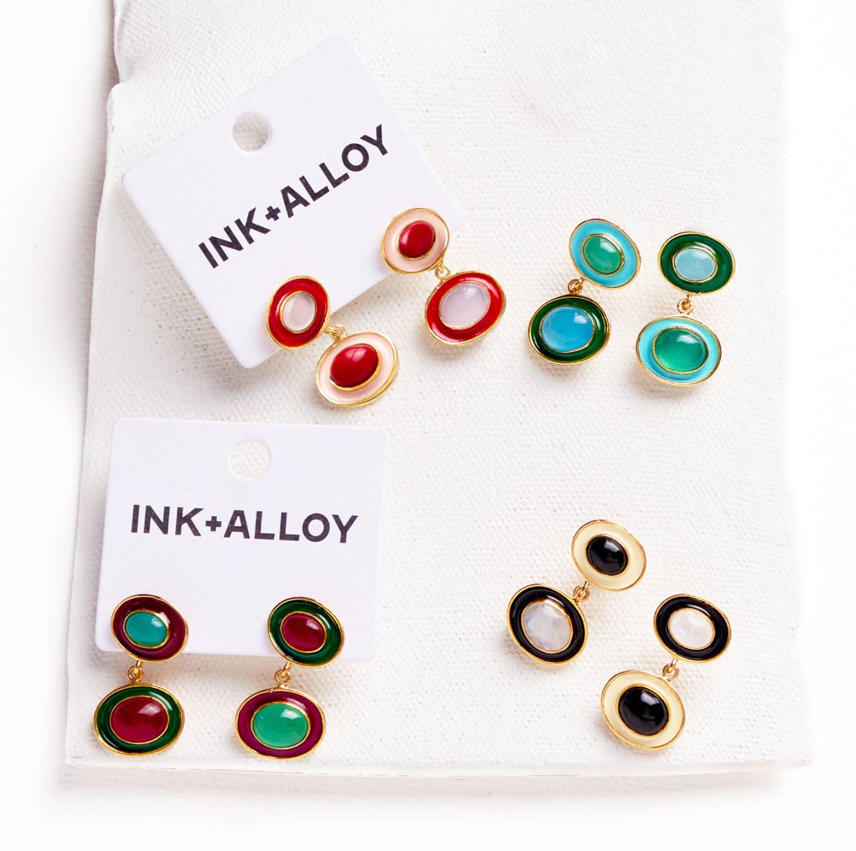 Four pairs of oval drop earrings displayed on white card hangers labeled INK+ALLOY, arranged on a white textured fabric background. The earrings feature gold-tone metal settings with oval stones in red, green, turquoise blue, and black.