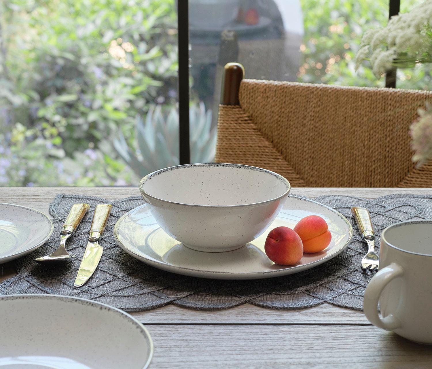 A ceramic table setting with a white speckled bowl on a matching plate, styled on a gray textured placemat with gold-handled cutlery and apricots, set in a bright dining area.