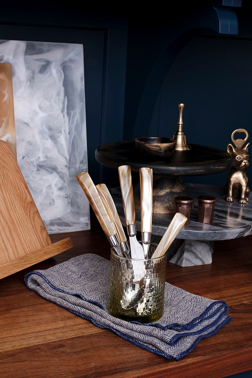 A styled kitchen or bar scene with a wooden cutting board on a stand, a glass jar holding several metal and light-colored utensils on a blue towel, a dark marble tiered stand, and decorative brass and gold objects on a wooden surface against a dark blue background.