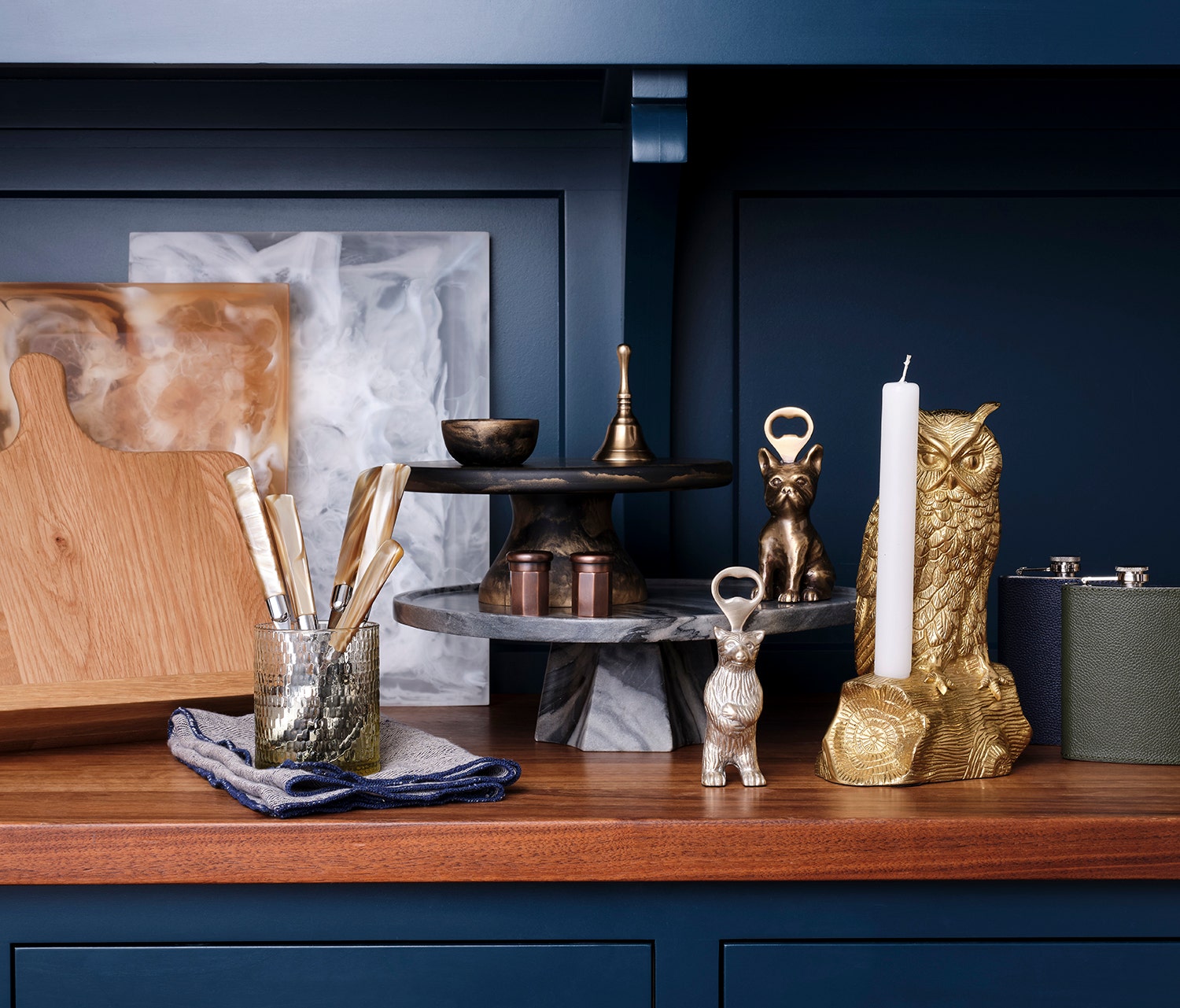 A styled kitchen countertop and shelf display featuring a gold owl-shaped candle holder, a white pillar candle, a small metallic deer figurine, a two-tiered marble and wood stand, a glass jar of utensils, and a wooden cutting board against a dark navy blue paneled background.