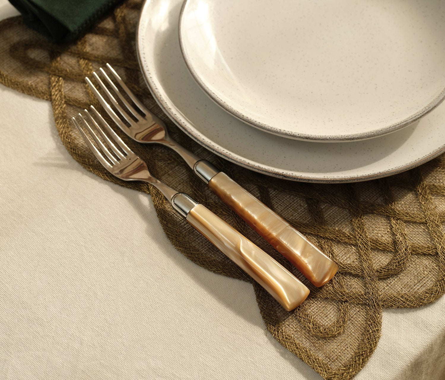 Two forks with polished silver-toned metal tines and cream-colored mother-of-pearl handles, arranged on a brown woven placemat next to stacked white ceramic plates on a white tablecloth.
