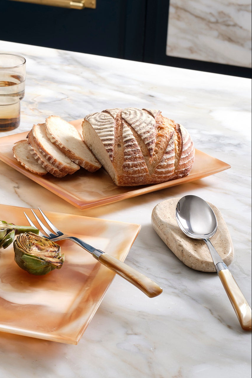 A styled dining table scene with a large rectangular peach-toned serving platter holding grilled artichokes and lemon wedges, a smaller matching platter with sliced bread, white plates, two glasses of water, and a spoon on a marble surface.