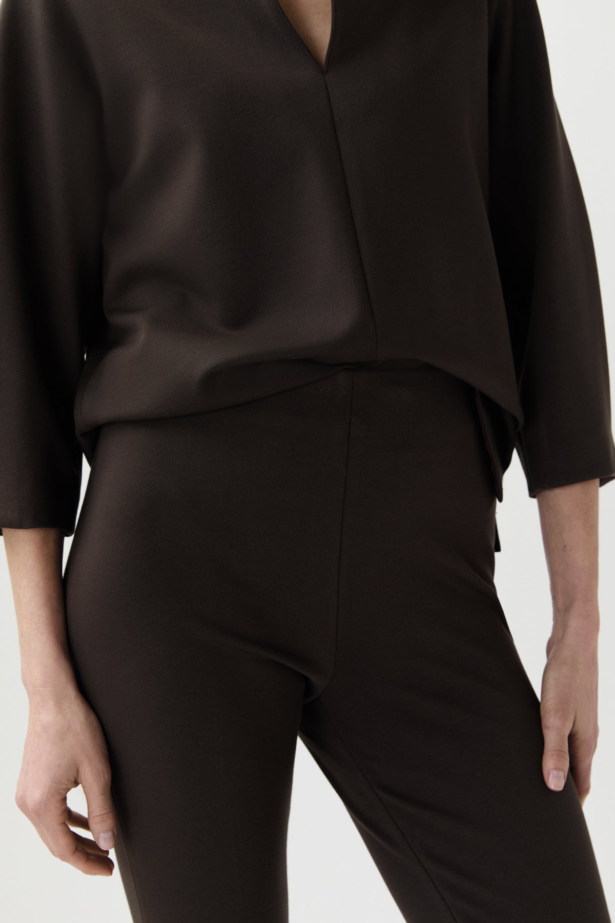 A close-up of a model wearing a dark brown satin long-sleeve top and matching dark brown trousers against a light gray background.