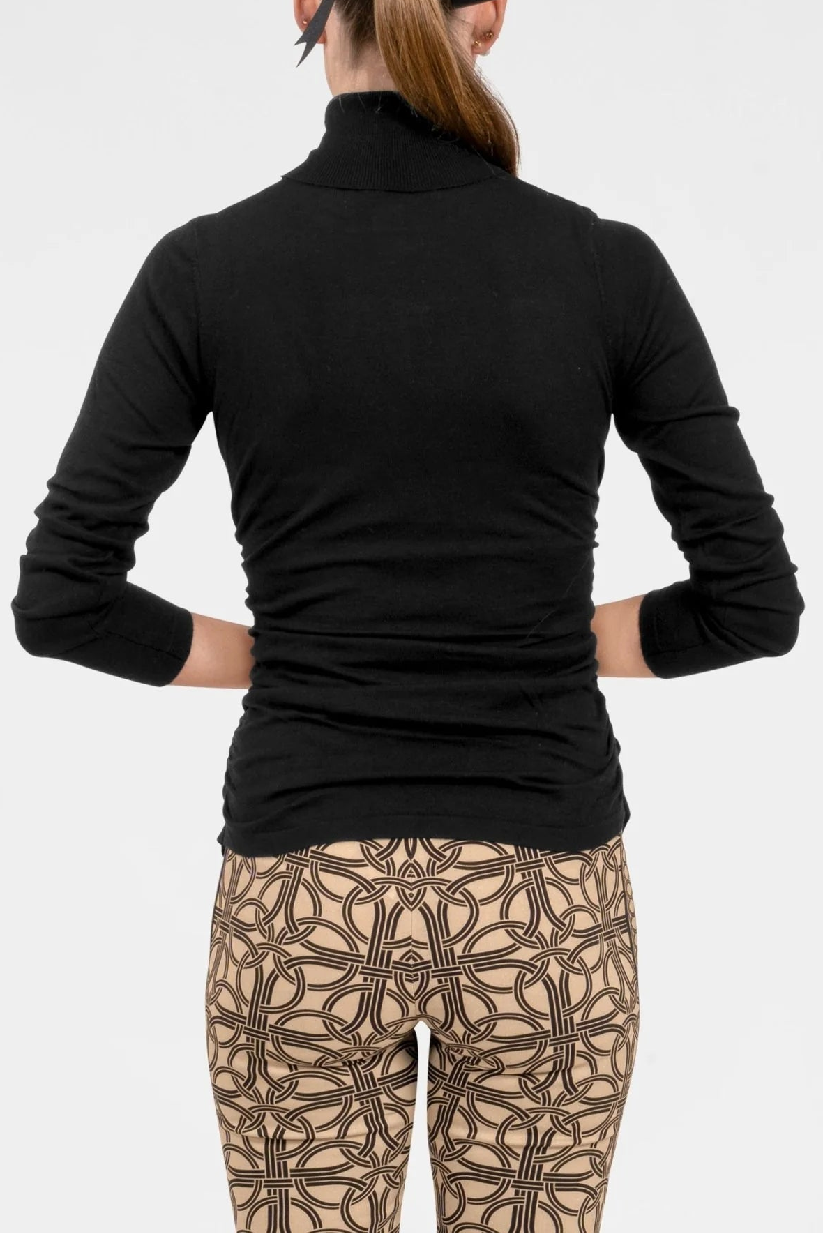 A woman shown from the back wearing a fitted black long-sleeve mock neck top and patterned beige leggings with a black bow in her hair, against a light gray studio background.
