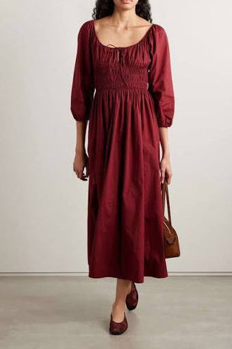 A woman models a long-sleeved midi dress in deep burgundy with a smocked bodice and square neckline, paired with brown flats and carrying a brown handbag against a neutral studio background.