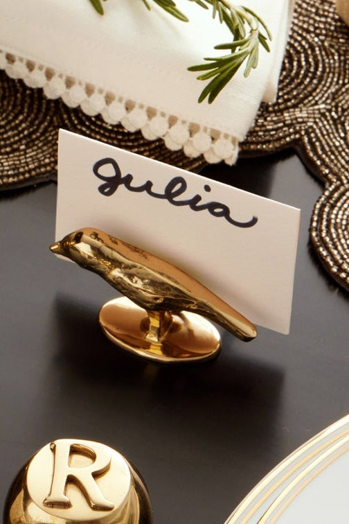 A gold bird-shaped place card holder on a dark table, holding a white card with the name 'Gulia' in black script. The scene includes decorative placemats, a white plate, and other gold table accessories.