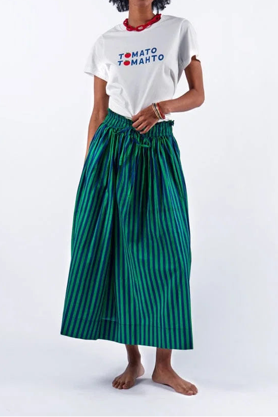 A woman with an afro hairstyle stands barefoot against a white studio background, wearing a white short-sleeve T-shirt with 'TOMATO TOMATO' printed on the chest in blue and red, paired with a high-waisted green pleated skirt featuring thin dark vertical stripes.