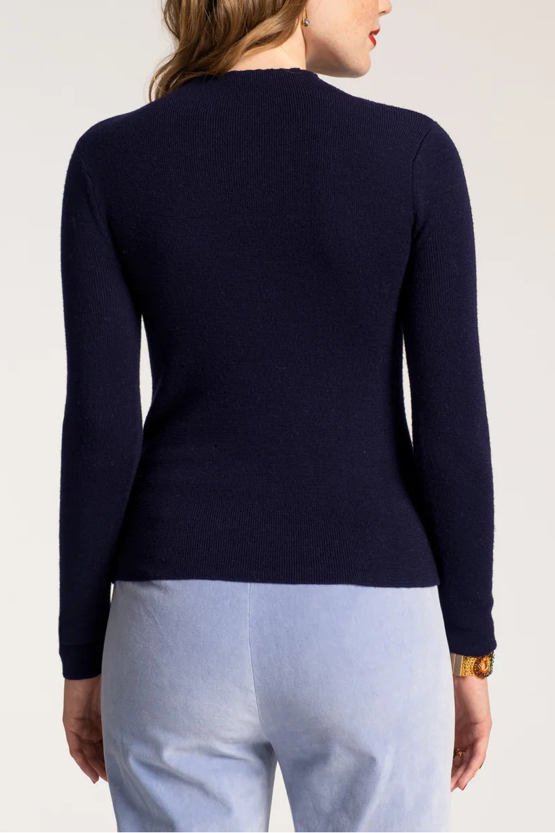 A woman shown from the back wearing a fitted long-sleeve knit top in dark navy blue and light blue pants, photographed against a plain light gray studio background.