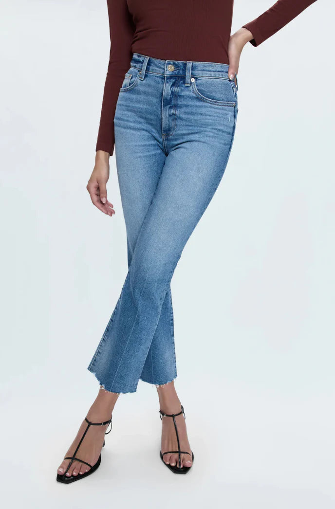 A model wearing high-waisted, cropped blue denim jeans with a subtle flare at the hem, paired with a dark brown long-sleeve top and black strappy heels against a light gray studio background.