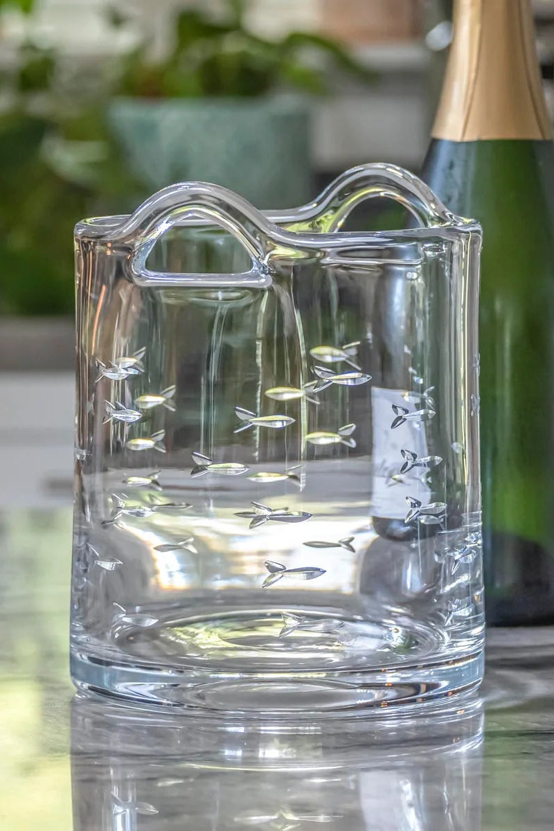 Clear glass ice bucket with engraved design on a reflective surface, with a bottle and glasses in the background.