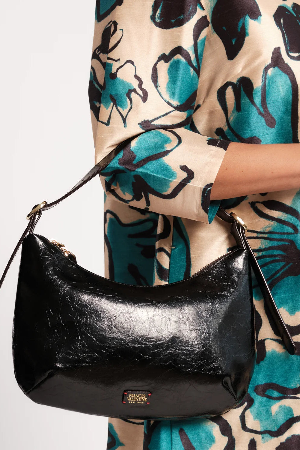 A black crescent-shaped shoulder bag with a glossy crinkled finish, worn by a model wearing a beige and teal floral dress. The bag features a small gold-tone brand plaque at the bottom center reading 'MARC FISHER NEW YORK'.